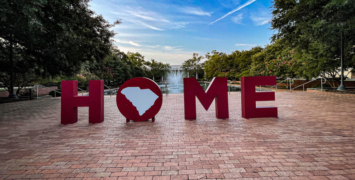 large garnet letters spelling out home on top of a brick walk way in front of the Cooper Library fountain with a blue ski