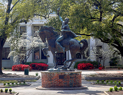 Metal Torchbearer statue on a brick base in front of Wardlaw college, surrounded by pathways and flowers. 