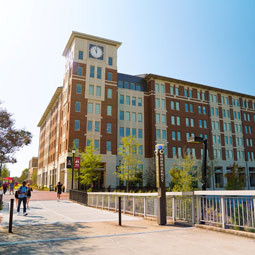 Students walk across a pedestrian bridge toward a campus village residence hall on a sunny day.