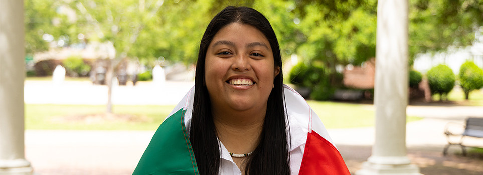 Student smiling while wearing a flag draped over her shoulders, standing in a shaded outdoor area with white columns and greenery in the background.