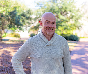 a man standing outside with a tan sweater on smiling. 