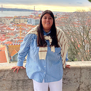 Student standing in front of a scenic overlook with red-roofed buildings and a suspension bridge in the distance, wearing a striped shirt and a sweater draped over her shoulders.