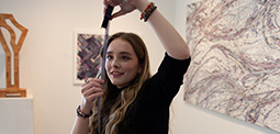 A woman with long hair holding a tool, standing in an art gallery with abstract paintings.