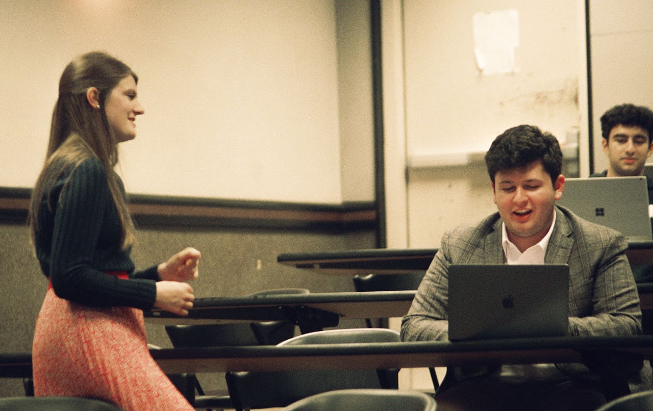 a photo of a person talking in a classroom with one student featured on a laptop. 