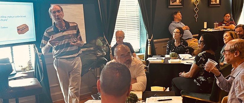 A man stands and speaks to a seated group during a workshop or meeting in a warmly lit room with dark walls, large windows, and modern hanging lights. Participants sit at round tables with papers and drinks as they listen.