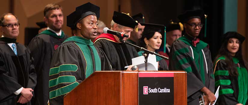 Speaker in academic regalia speaks into a microphone at a podium