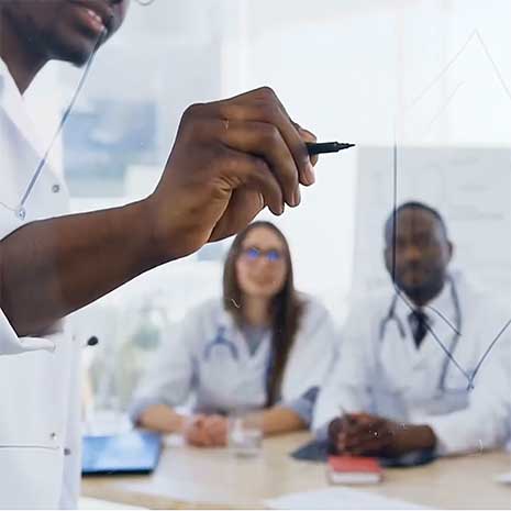 Doctor in white coat writes on clear board while students watch from table.