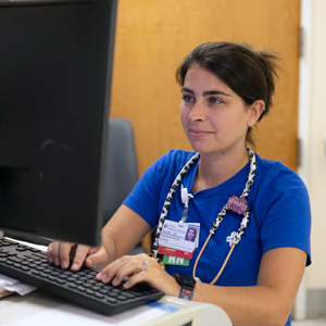 woman in scrubs with a stethoscope around her neck works at a computer station