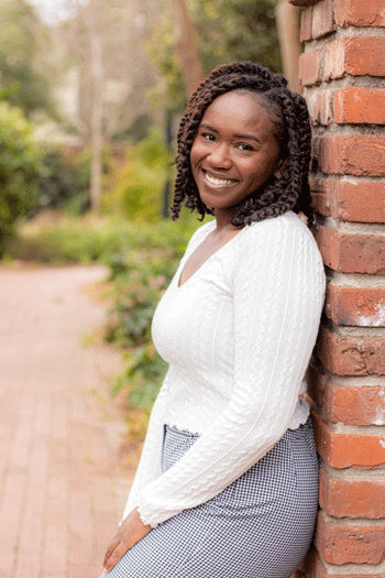 Taylor Jennings-Brown wearing a white sweater leans against brick gate on University of South Carolina's Horseshoe.