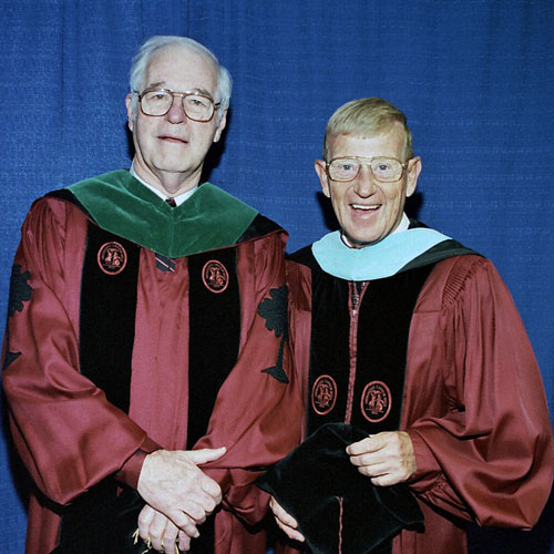 two men wearing academic regalia stand in front of a blue background